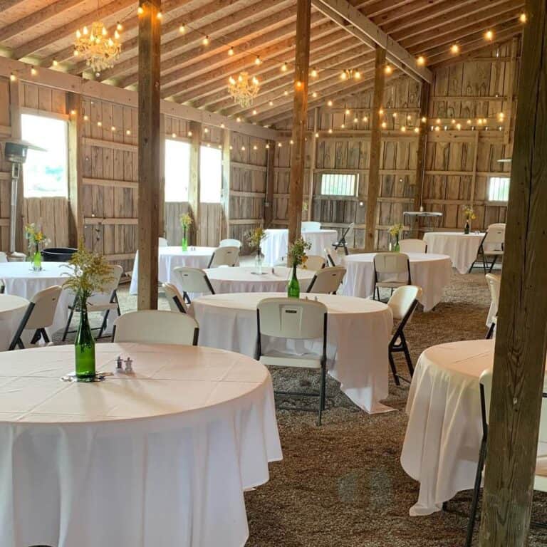 Round tables with white linens and chairs set up under string lights inside the barn at Highland Oaks in Palmyra, Indiana
