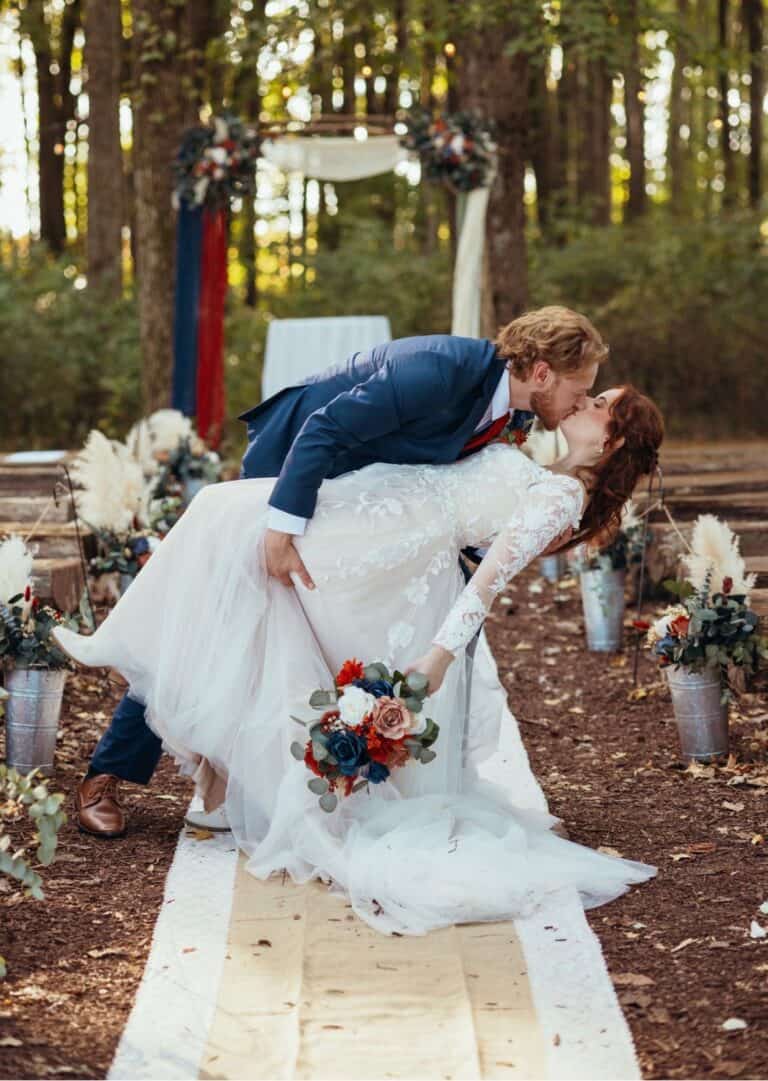 Couple sharing a dip kiss on the wooden aisle of the forest ceremony site at Highland Oaks in Palmyra, Indiana, with the floral arch and bouquets on stands behind them