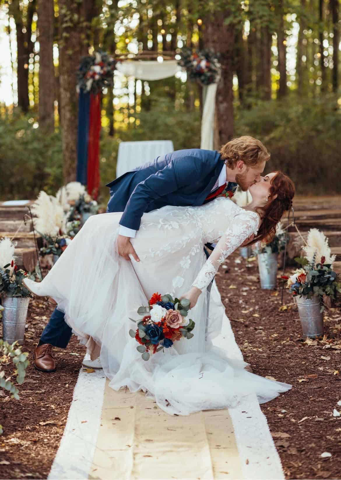 Couple sharing a dip kiss on the wooden aisle of the forest ceremony site at Highland Oaks in Palmyra, Indiana, with the floral arch and bouquets on stands behind them