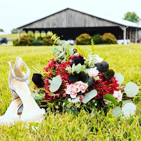 Bridal heels and a red and blush bouquet resting on the grass in front of the barn at Highland Oaks in Palmyra, Indiana