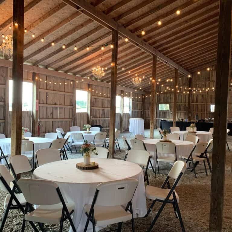 Barn reception at Highland Oaks in Palmyra, Indiana, with round tables in white linens, folding chairs, string lights and chandeliers under the wood beams