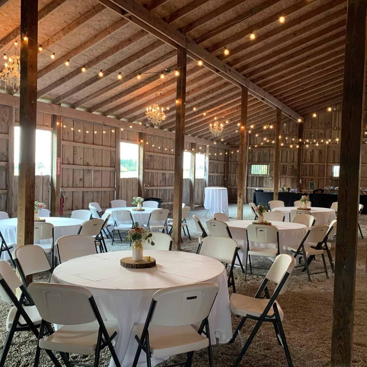 Barn reception at Highland Oaks in Palmyra, Indiana, with round tables in white linens, folding chairs, string lights and chandeliers under the wood beams