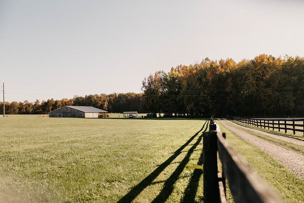 Long fence shadows across the field at Highland Oaks in Palmyra, Indiana, with the barn and oak treeline in late day light